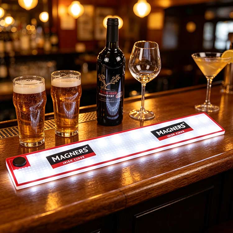 Battery operated illuminated commercial bar runner laid on warm wooden Irish pub bar counter, Magners branded glowing bar spill mat paired with cider pints, wine bottle and cocktail glasses for classic pub decor