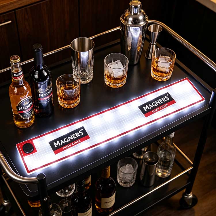 Waterproof commercial LED bar runner laid on silver trimmed luxury black metal home bar cart, Magners glowing illuminated bar spill mat styled with whiskey tumblers, cocktail shaker, jigger and full cider wine bottles for premium home bar decor