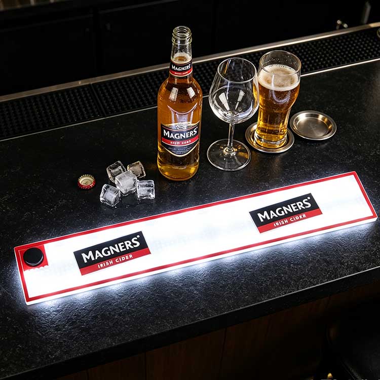 Waterproof commercial LED bar runner on dark stone pub bar counter, glowing Magners logo bar spill mat styled with full cider bottle, pint glass filled with cider, ice cubes and bottle cap for daily pub service