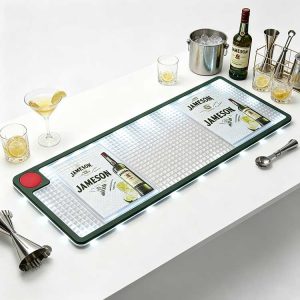 White background studio photo of branded LED bar mat printed with Jameson whiskey logo, waterproof non-slip spill-proof illuminated bar drip runner with glowing LED edge, arranged with cocktail glasses, whiskey bottle, metal jigger and ice bucket for bar counter use
